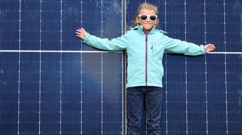 Mädchen mit Sonnenbrille steht vor einem Solarpanel die Hände ausgebreitet